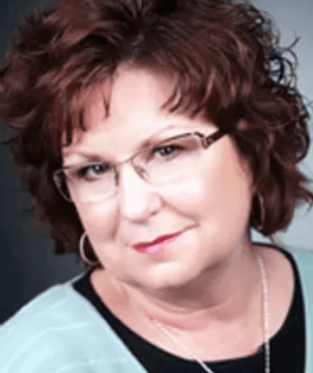 Person with curly hair and glasses, wearing a light-colored top, smiling slightly at the camera.