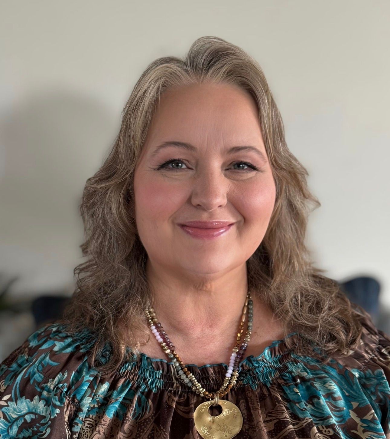 Smiling person with wavy hair, wearing a patterned top and beaded necklace with pendant.